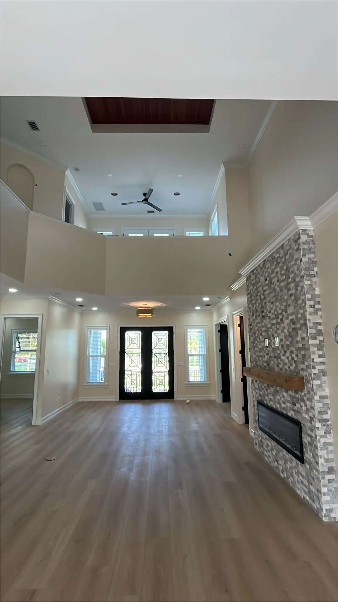 A spacious room with light-colored laminate flooring, a stone-faced fireplace with a wooden mantel, a black double door with decorative grilles, and a double-height ceiling with recessed lighting and a dark wooden panel is shown from a wide angle.