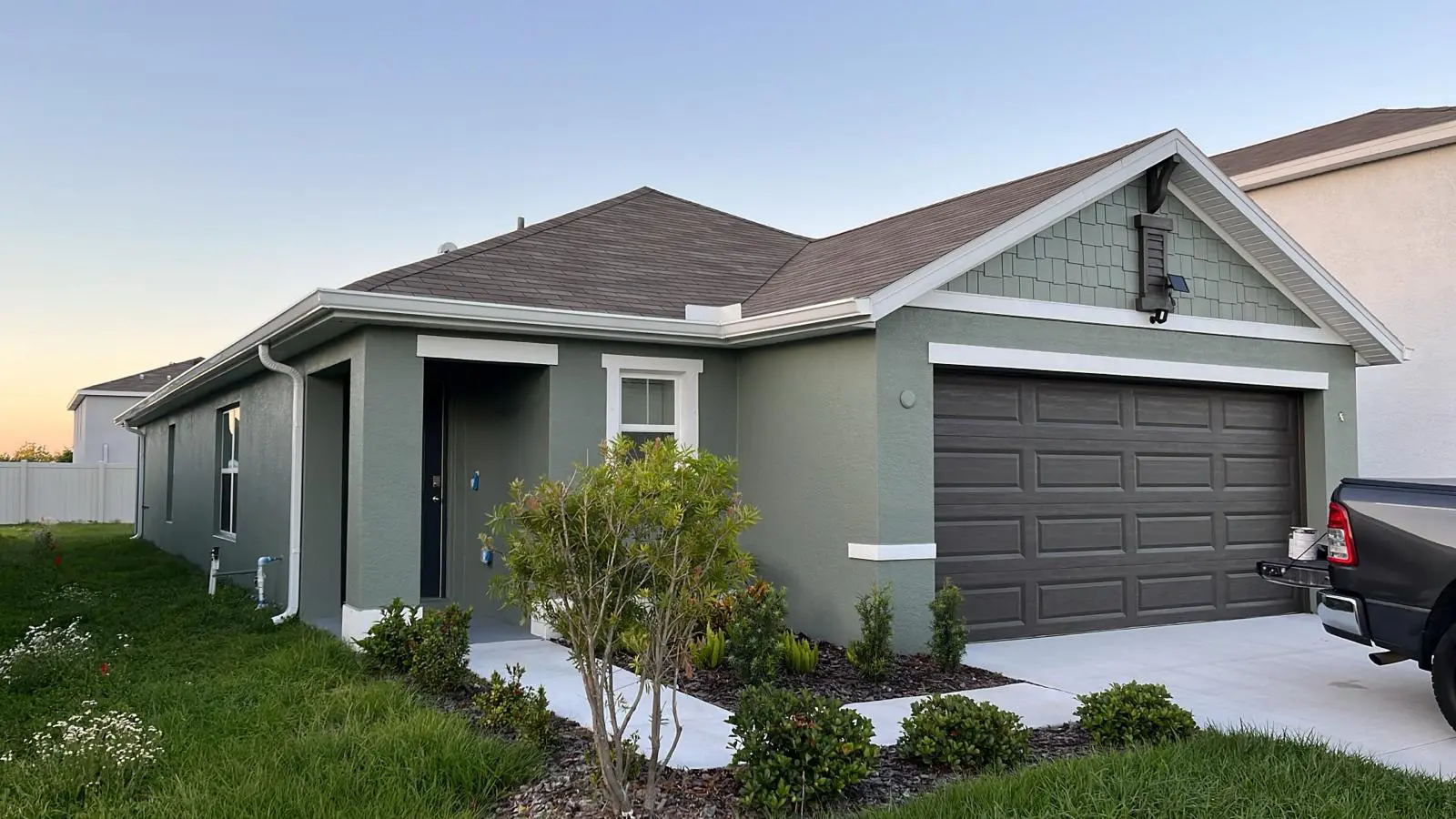 A freshly painted gray house with a darker gray garage door and a brown shingle roof is bathed in the warm light of the setting sun, with a neatly landscaped yard and a dark truck parked in the driveway.