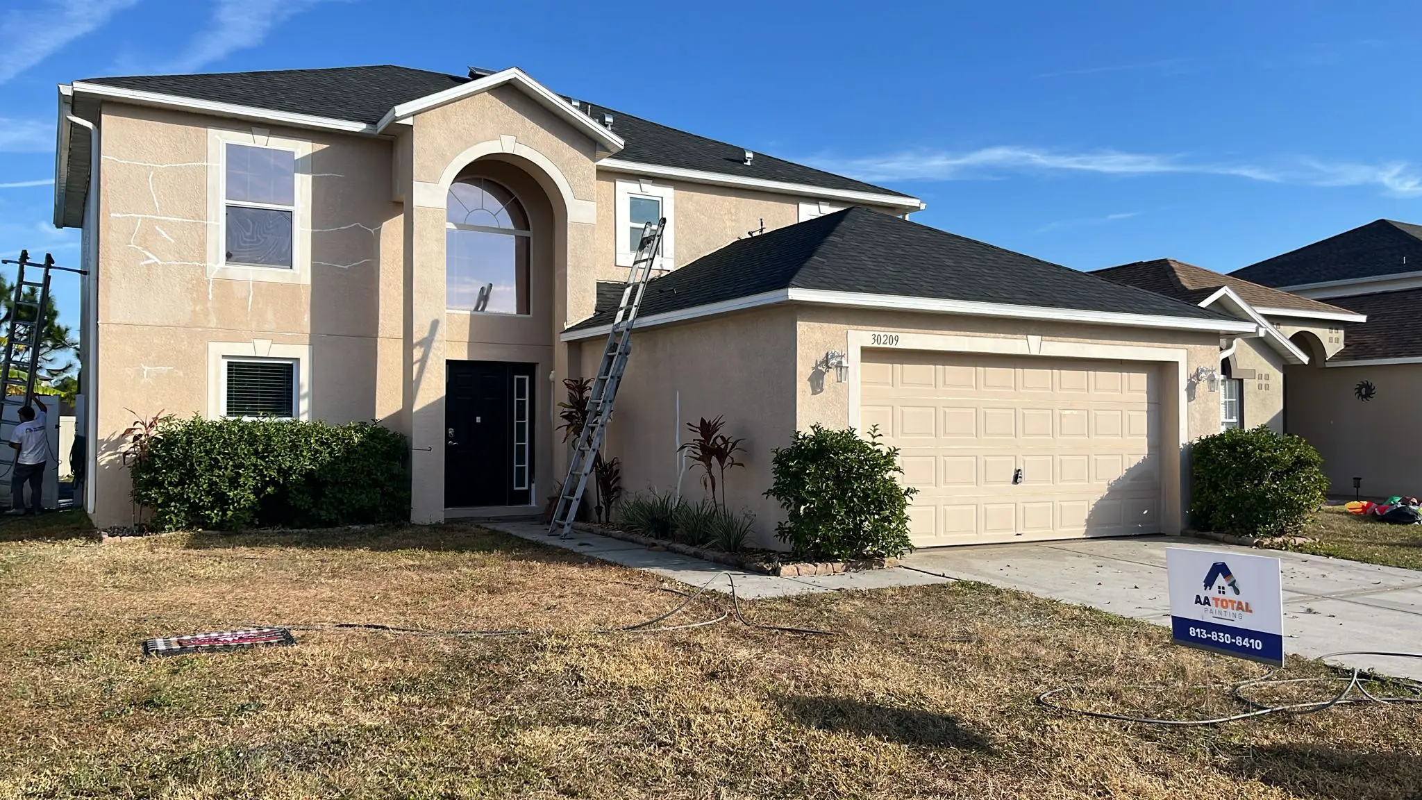A two-story beige house with a dark gray roof and a light beige garage door is being prepared for painting, with ladders, tarps, and an "AA TOTAL" sign visible in the yard under a sunny sky.