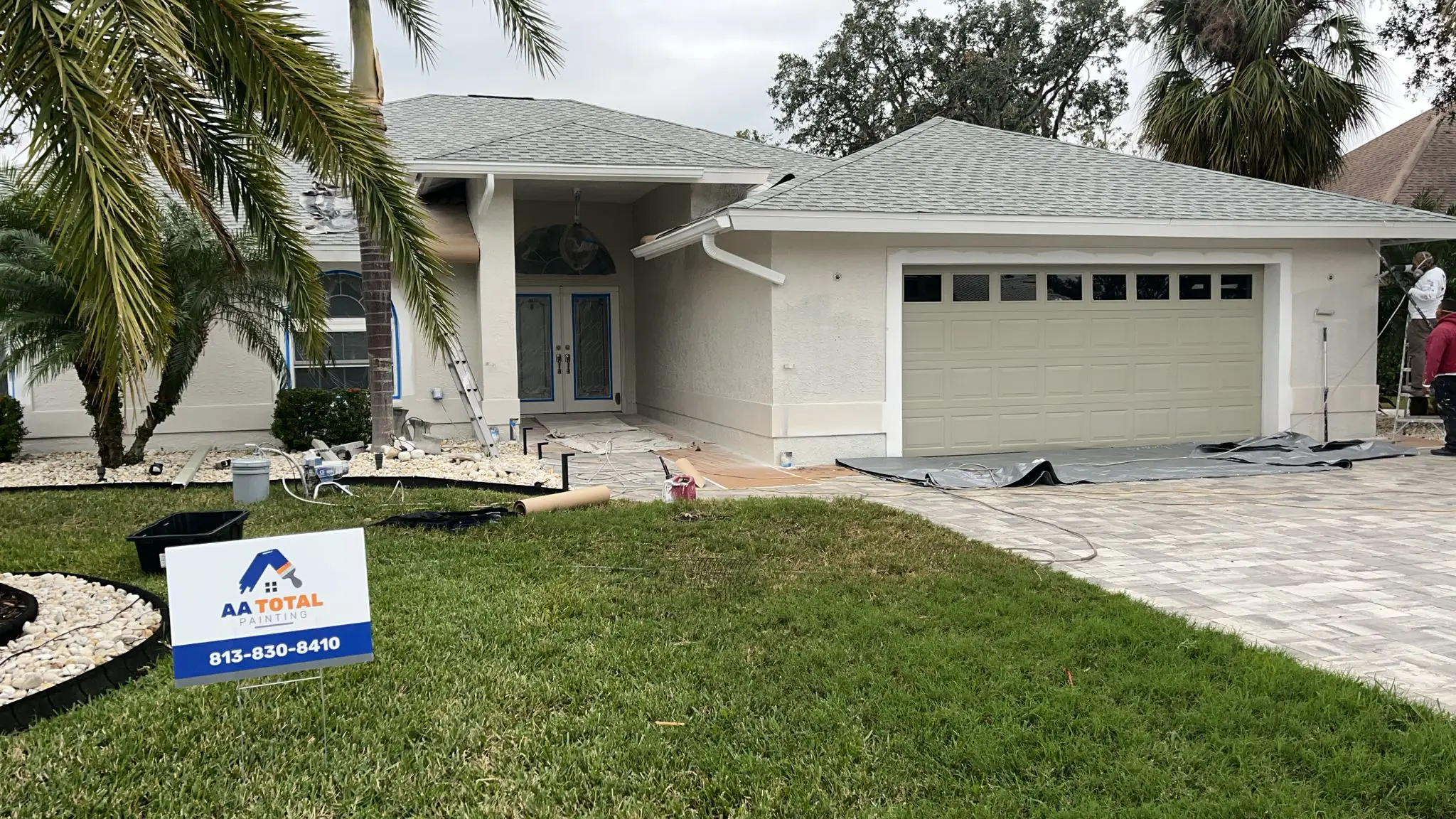 A light-colored house with a gray garage door and a gray shingle roof is undergoing painting, with ladders, tarps, and an "AA TOTAL PAINTING" sign visible in the yard, under a cloudy sky.