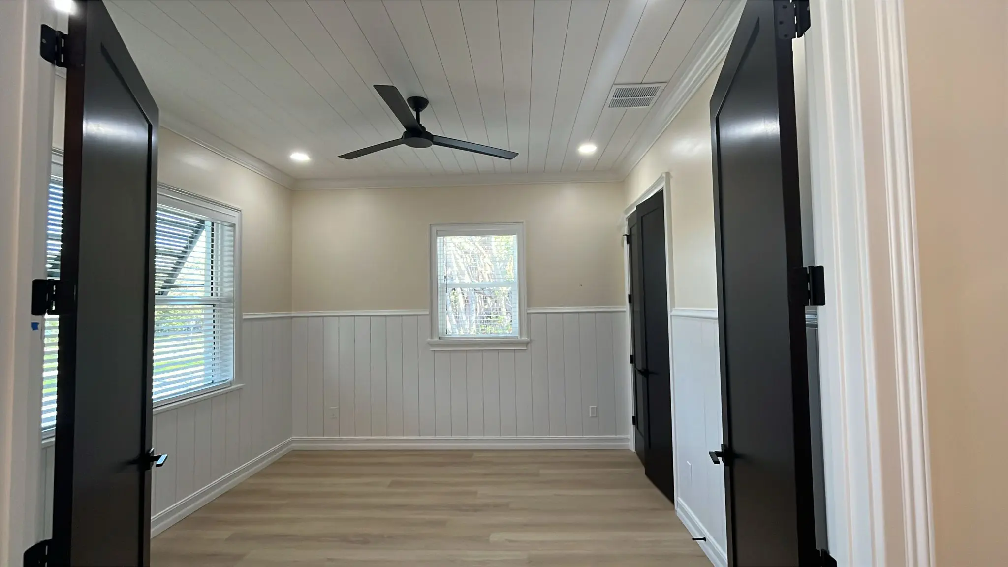A bright room with white shiplap walls, light wood laminate flooring, two windows with blinds, recessed lighting, and a black ceiling fan is viewed from a corner angle. A bright room with white shiplap walls, light wood laminate flooring, two windows with blinds, recessed lighting, and a black ceiling fan is viewed from a corner angle.