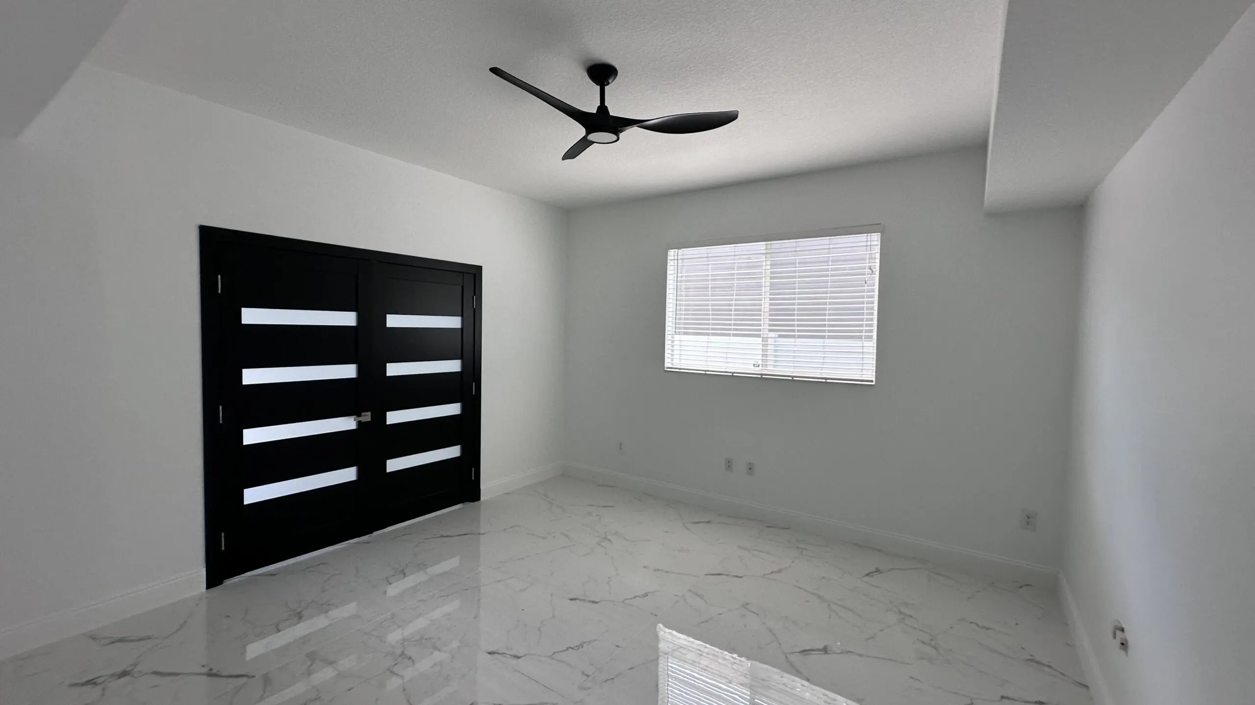 A bright room features a black ceiling fan, white walls, marble-patterned tile flooring, a window with blinds, and a black double door with white horizontal stripes.