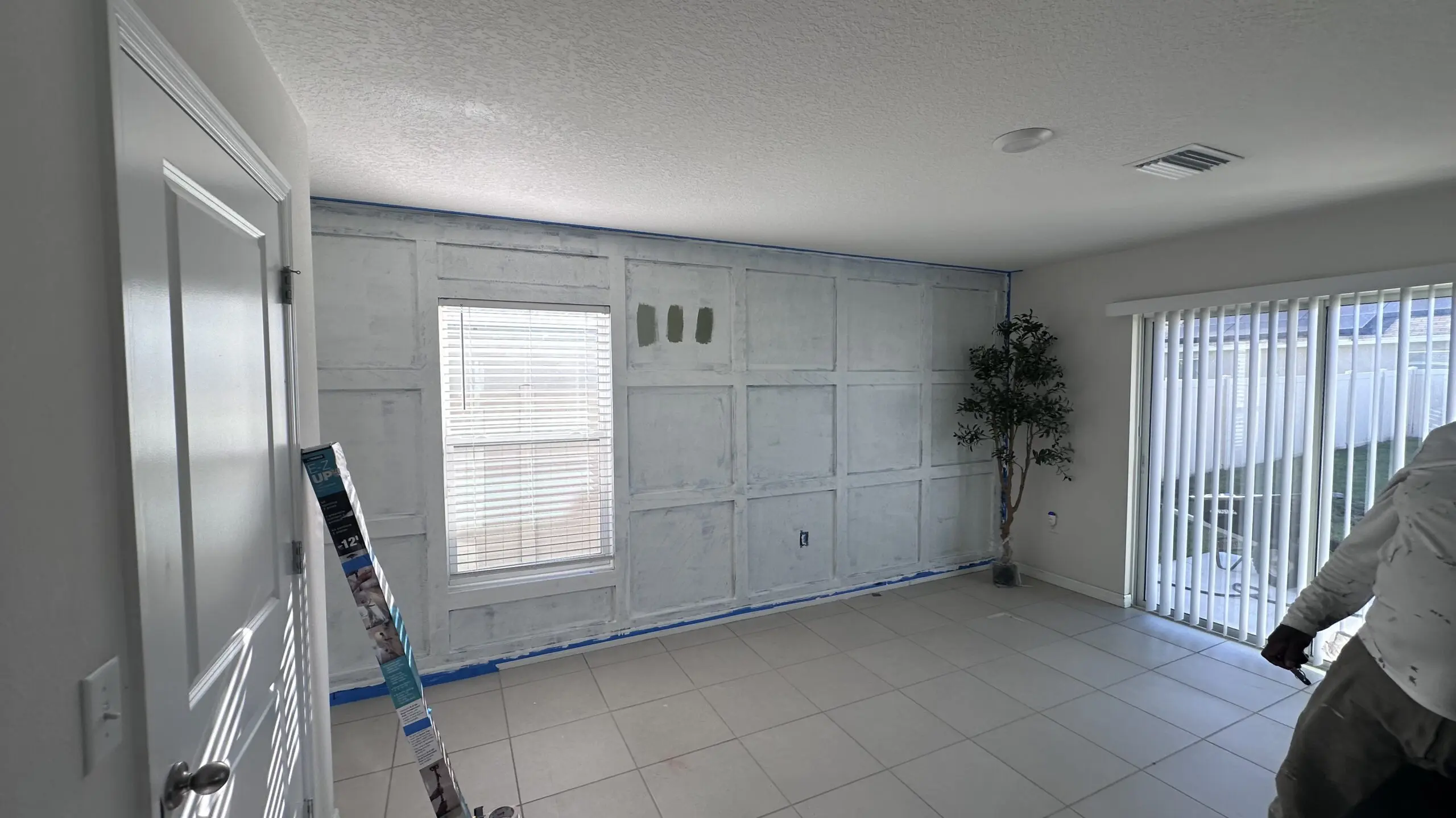 An interior room with white textured walls and a white textured ceiling features a wall with panel molding that is partially painted in a lighter shade, a window with blinds, sliding glass doors leading outside, a potted plant, and a partially visible person in the corner. An interior room with white textured walls and a white textured ceiling features a wall with panel molding that is partially painted in a lighter shade, a window with blinds, sliding glass doors leading outside, a potted plant, and a partially visible person in the corner.