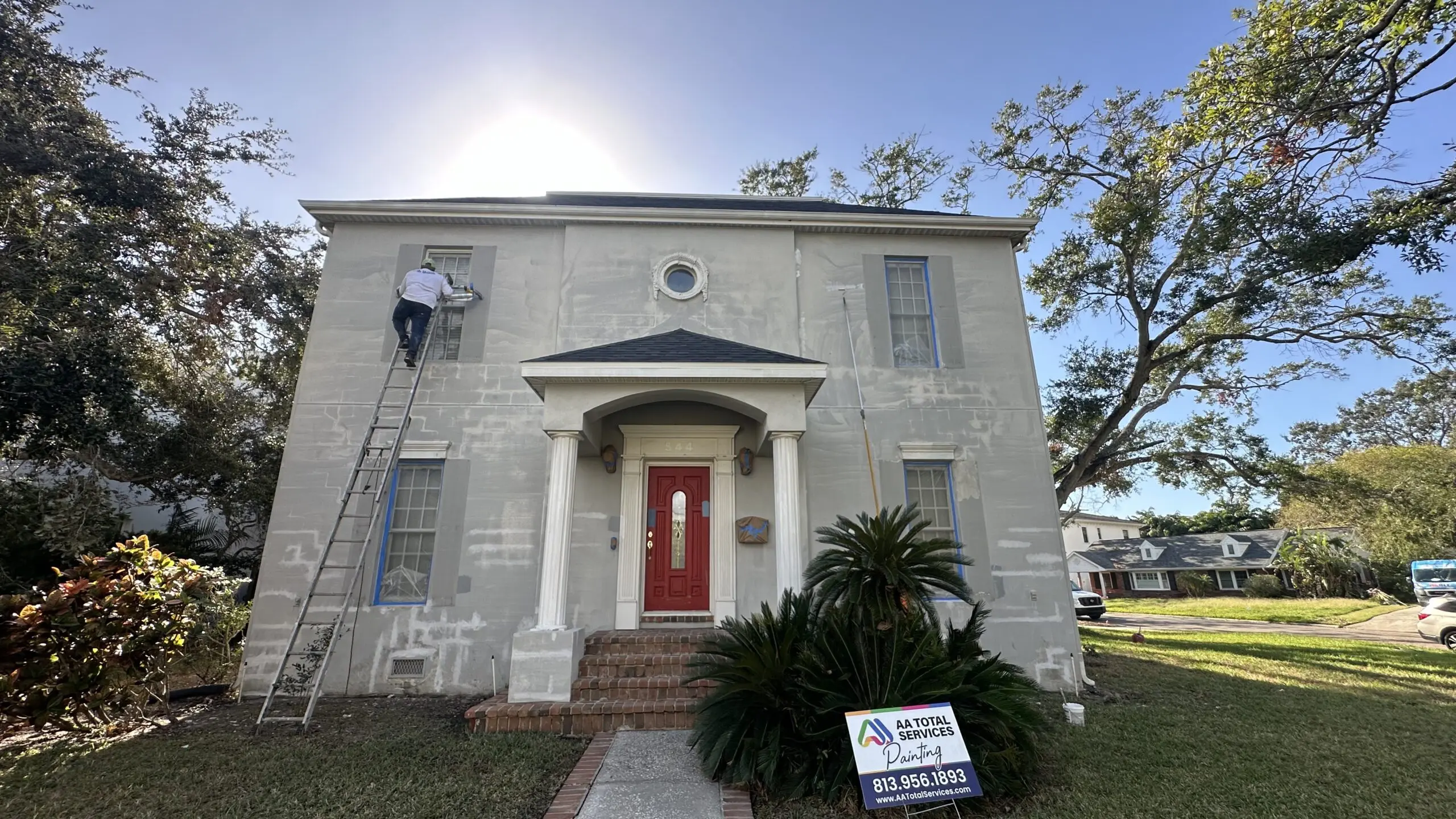 A painter on a ladder is working on the exterior of a light gray two-story house with a red front door, with an "AA TOTAL SERVICES Painting" sign in the front yard, under a bright sun.