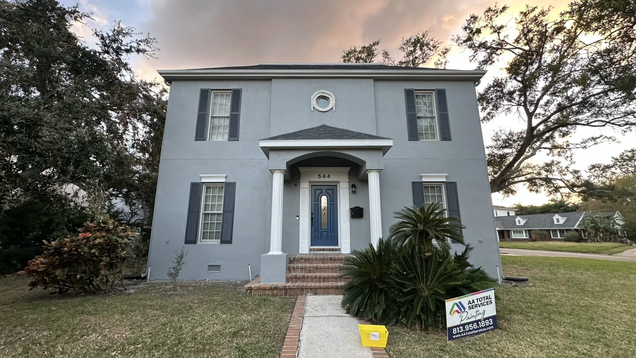 A freshly painted gray two-story house with dark blue shutters and a matching front door is centered in the frame, with an "AA TOTAL SERVICES Painting" sign and a yellow bucket in the front yard, under a dramatic sky at dusk.