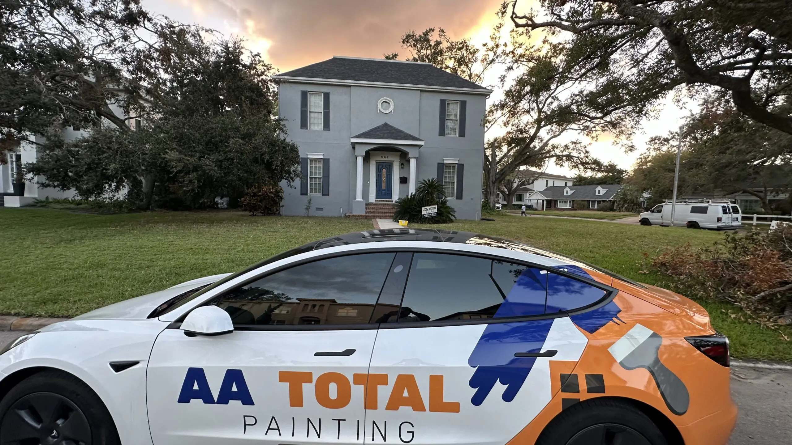 A white Tesla Model 3 with an "AA TOTAL PAINTING" logo is parked in front of a recently painted gray house with a dark gray roof, under a dramatic sky.