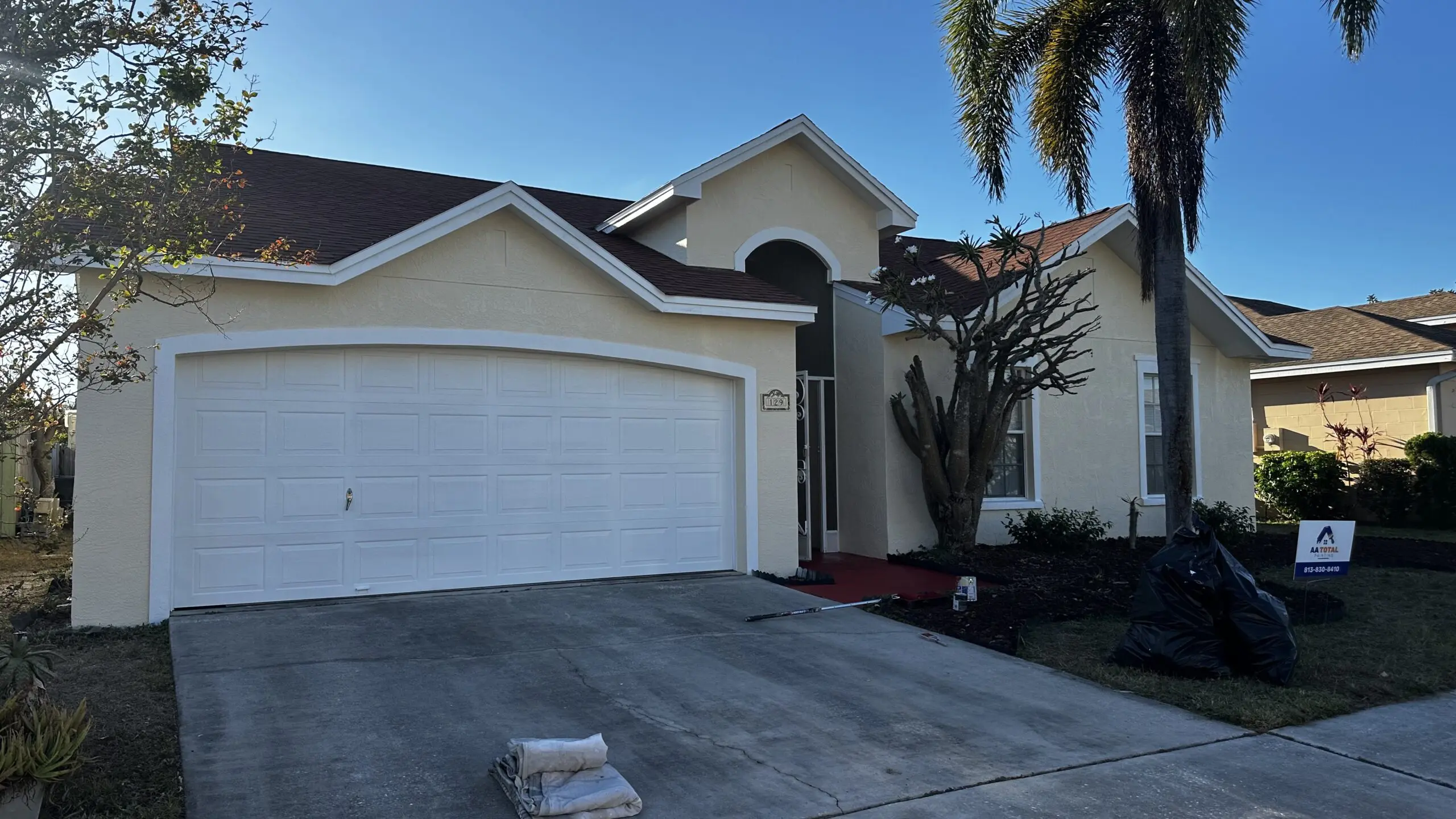 A freshly painted beige house with a white garage door and a brown roof is bathed in sunlight, with a palm tree casting a shadow and an "AA TOTAL" sign visible in the yard.