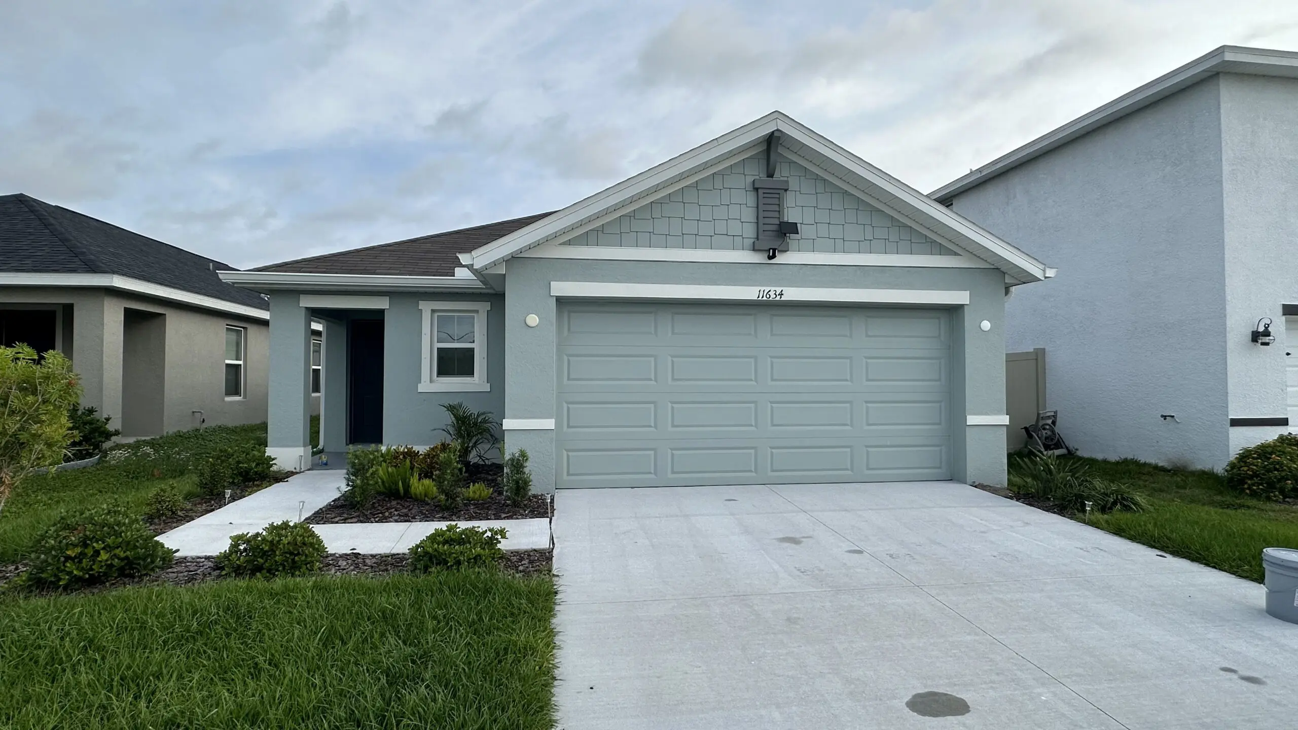 A light gray single-story house with a gray garage door and a dark gray roof is nestled between two other houses, with a narrow strip of lawn leading to the front door, under a cloudy sky.