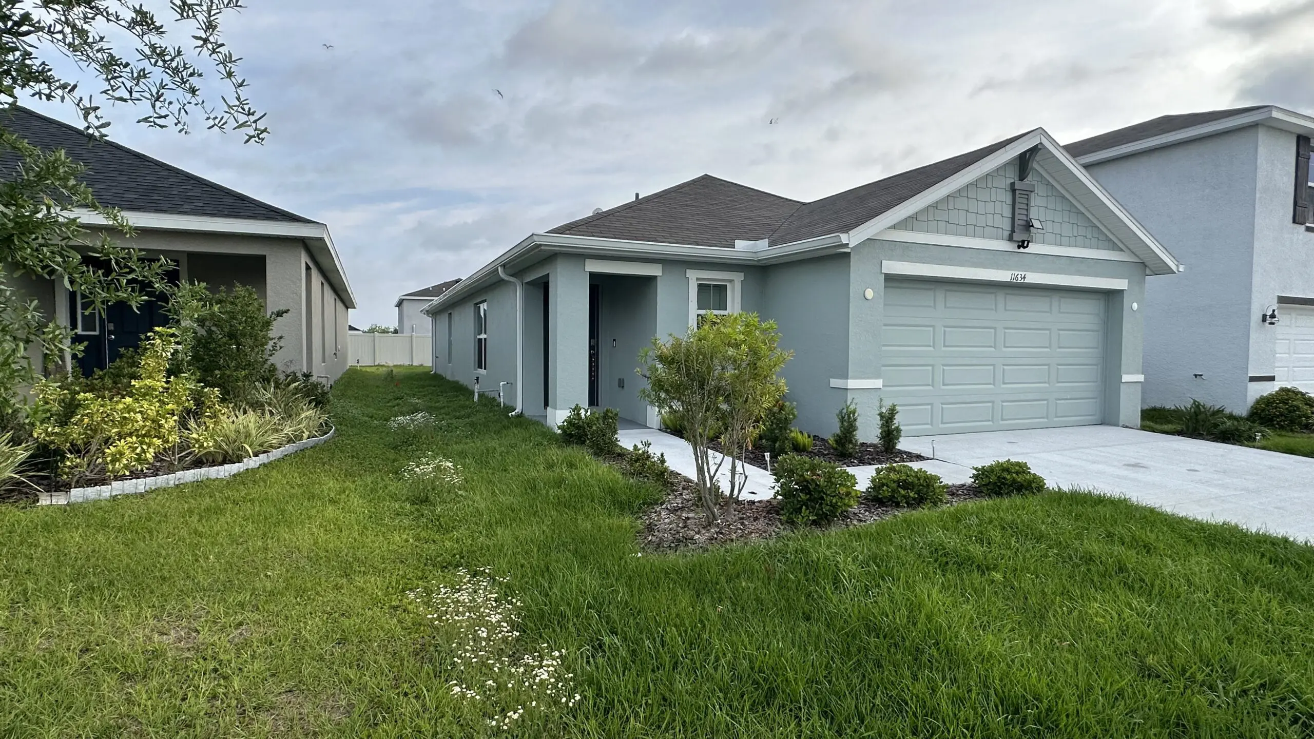 A light gray single-story house with a gray garage door and a dark gray roof is nestled between two other houses, with a narrow strip of lawn leading to the front door, under a cloudy sky.