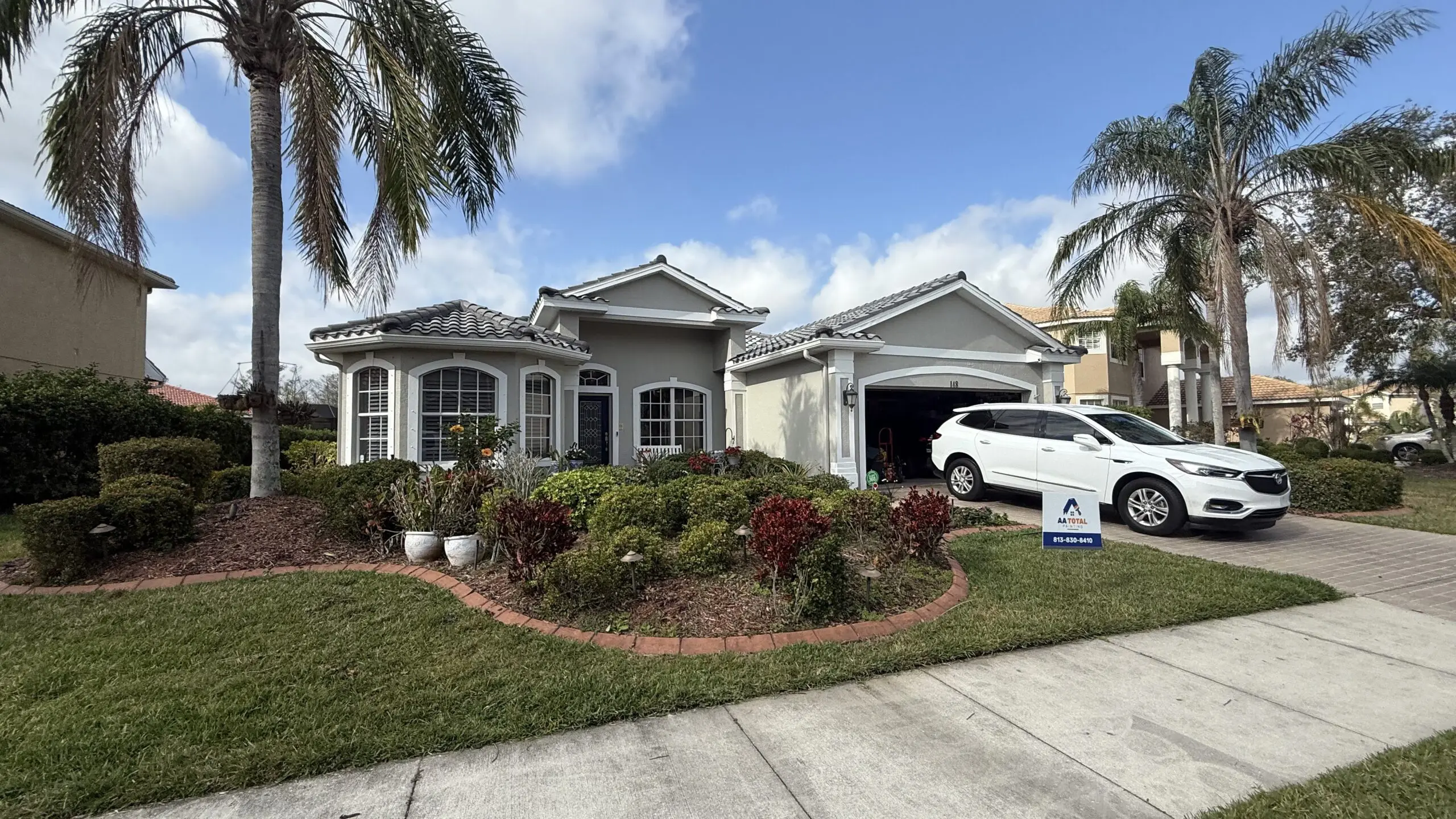 A gray house with a gray tile roof and arched windows is surrounded by lush landscaping, with a white SUV parked in the driveway and tall palm trees framing the scene under a partly cloudy sky.