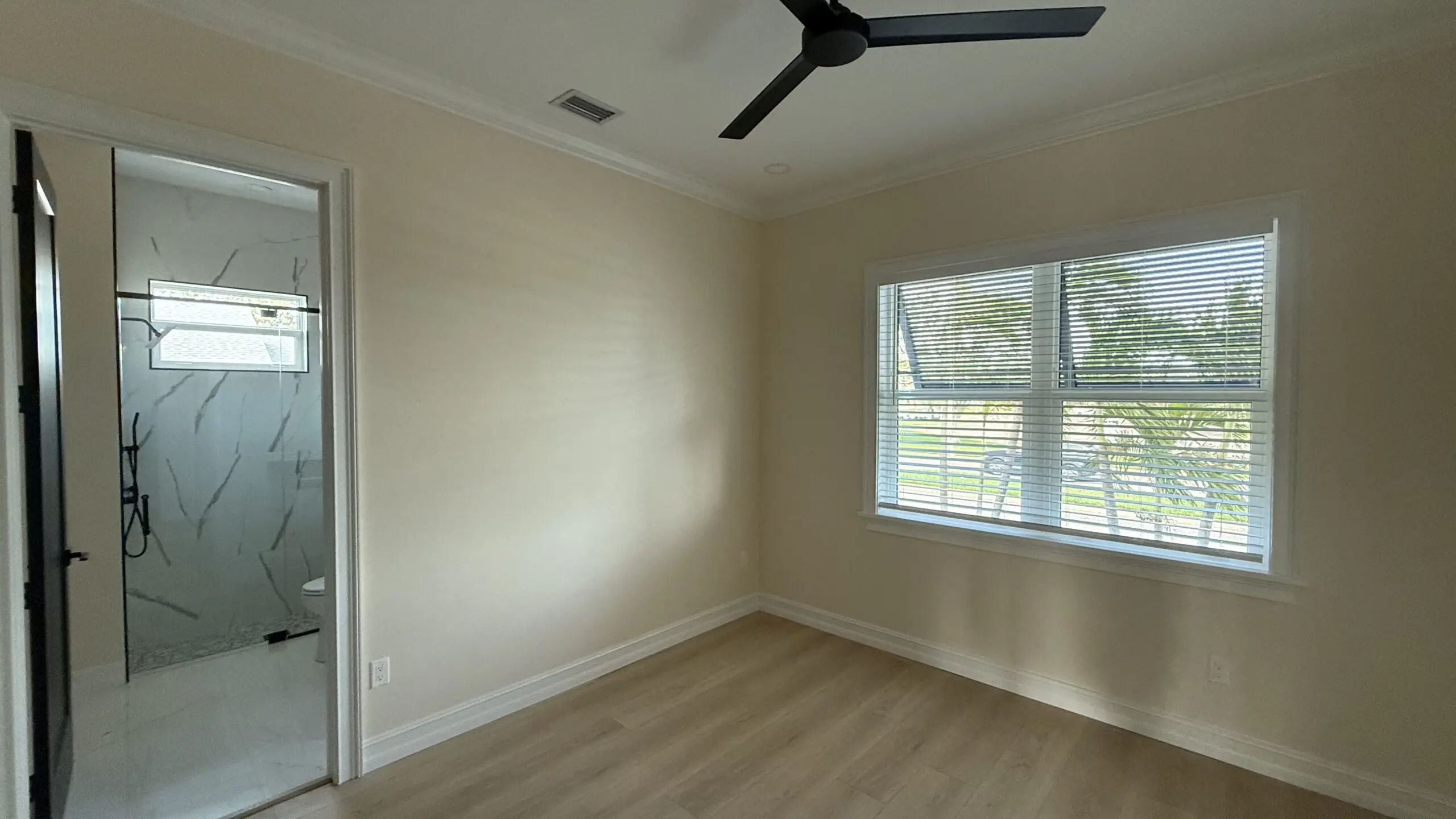 A bright room features a black ceiling fan, light-colored walls, wood laminate flooring, a window with blinds, and a doorway leading to a bathroom with a glass-enclosed shower. A bright room features a black ceiling fan, light-colored walls, wood laminate flooring, a window with blinds, and a doorway leading to a bathroom with a glass-enclosed shower.