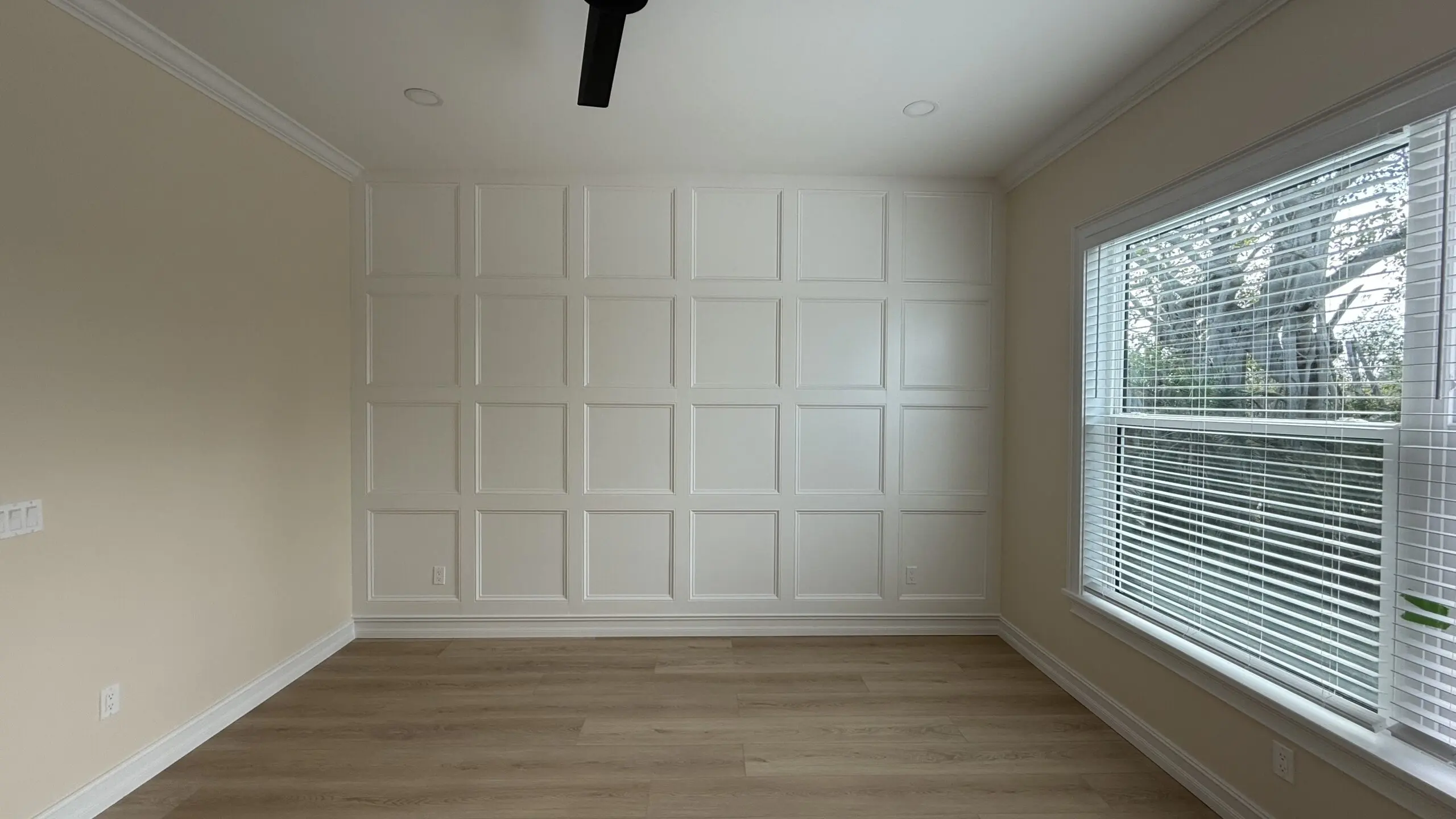 A room features a wall with white wainscoting panels in a grid pattern, light-colored walls, wood laminate flooring, a large window with blinds, and a black ceiling fan. A room features a wall with white wainscoting panels in a grid pattern, light-colored walls, wood laminate flooring, a large window with blinds, and a black ceiling fan.