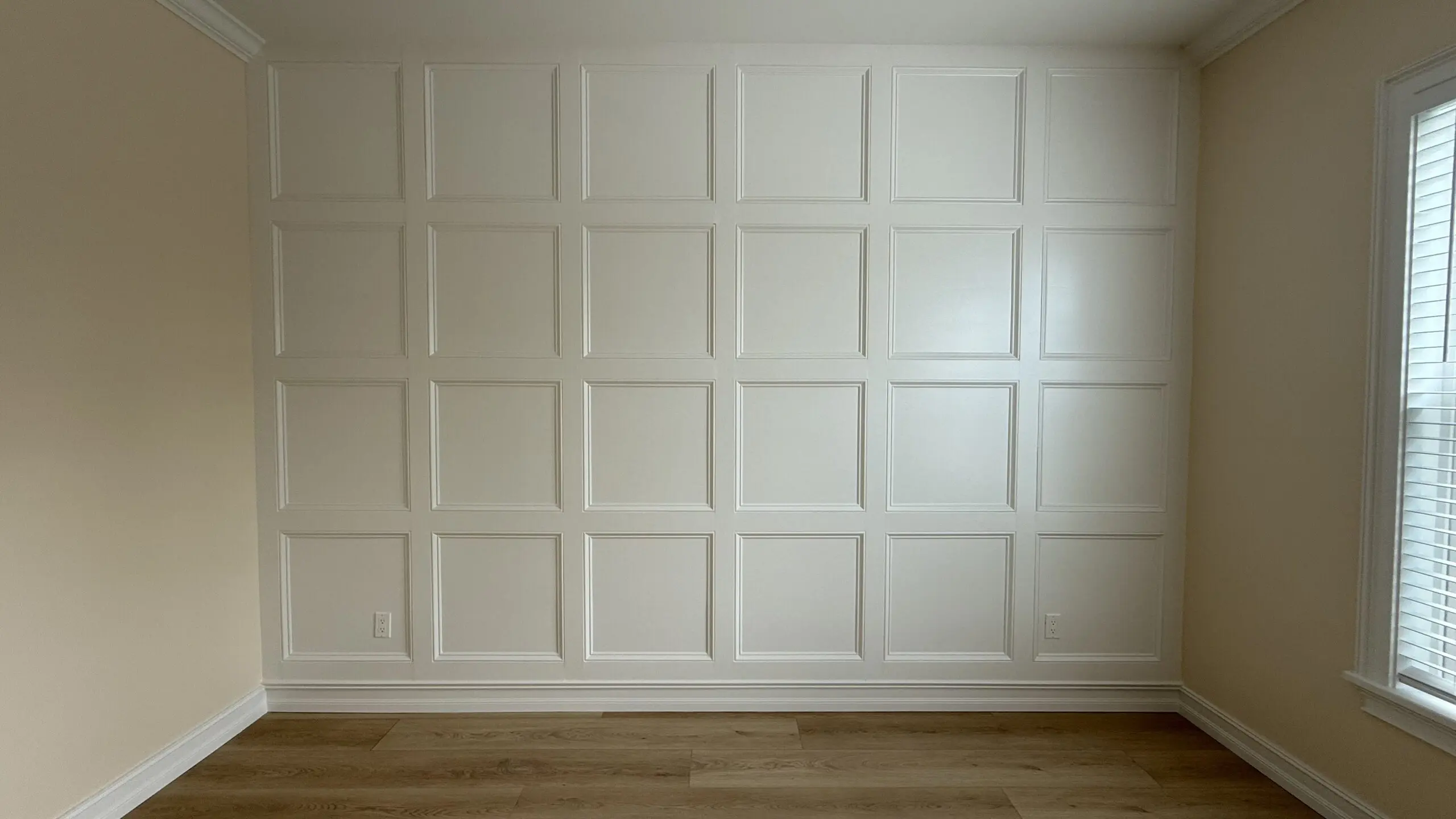A wall with white wainscoting panels in a grid pattern is featured in a room with light-colored walls, wood laminate flooring, and a window with blinds on the right. A wall with white wainscoting panels in a grid pattern is featured in a room with light-colored walls, wood laminate flooring, and a window with blinds on the right.