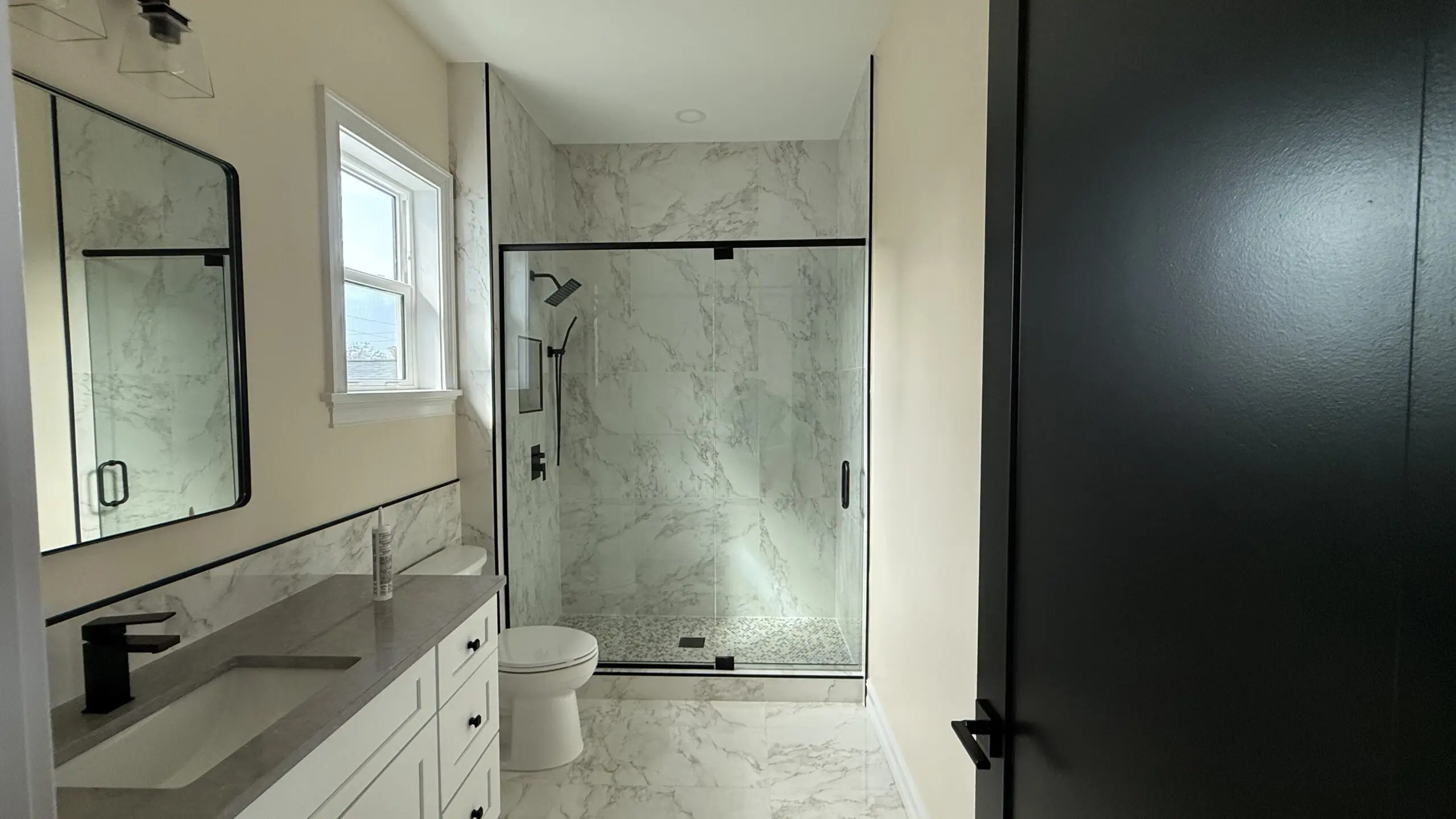 A modern bathroom features a glass-enclosed shower with marble-patterned walls and floor, a white toilet, a vanity with a gray countertop and double sinks, a framed mirror, a window, and a dark-colored door partially visible on the right. A modern bathroom features a glass-enclosed shower with marble-patterned walls and floor, a white toilet, a vanity with a gray countertop and double sinks, a framed mirror, a window, and a dark-colored door partially visible on the right.
