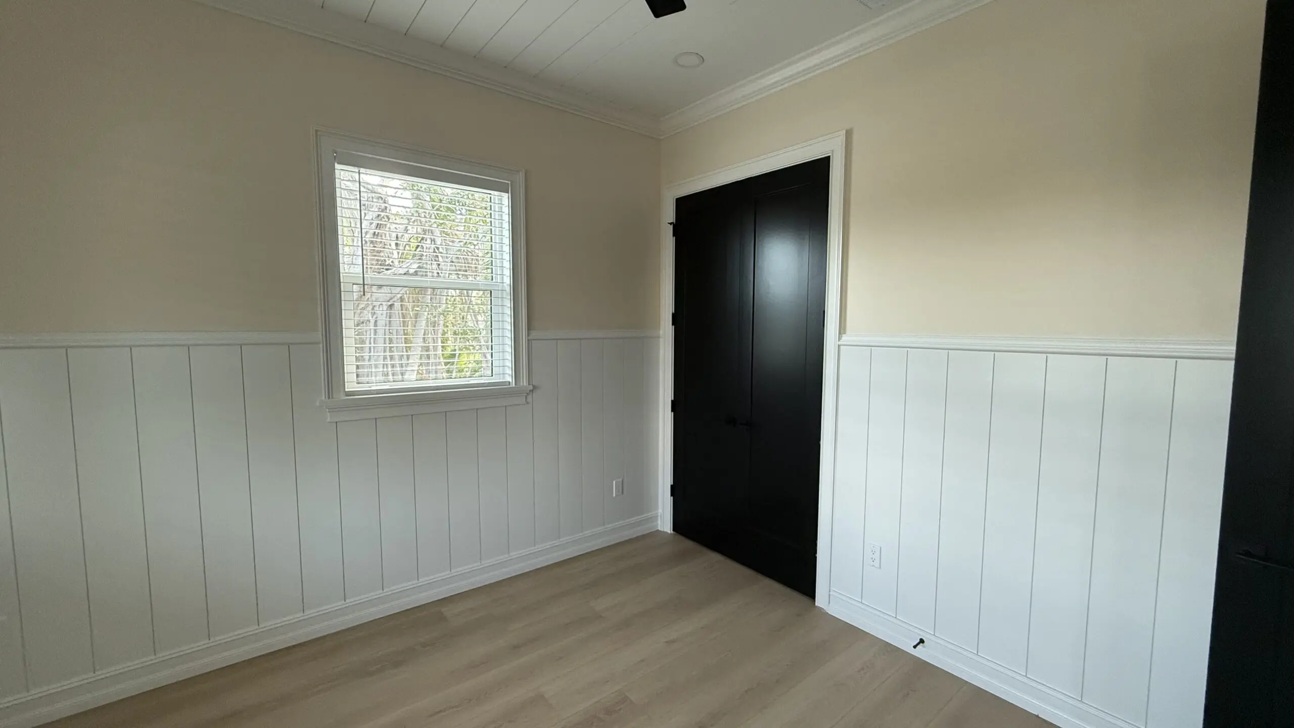 A small room with light-colored walls, white wainscoting, light wood laminate flooring, a window with blinds, and a dark-colored door is viewed from a corner angle. A small room with light-colored walls, white wainscoting, light wood laminate flooring, a window with blinds, and a dark-colored door is viewed from a corner angle.