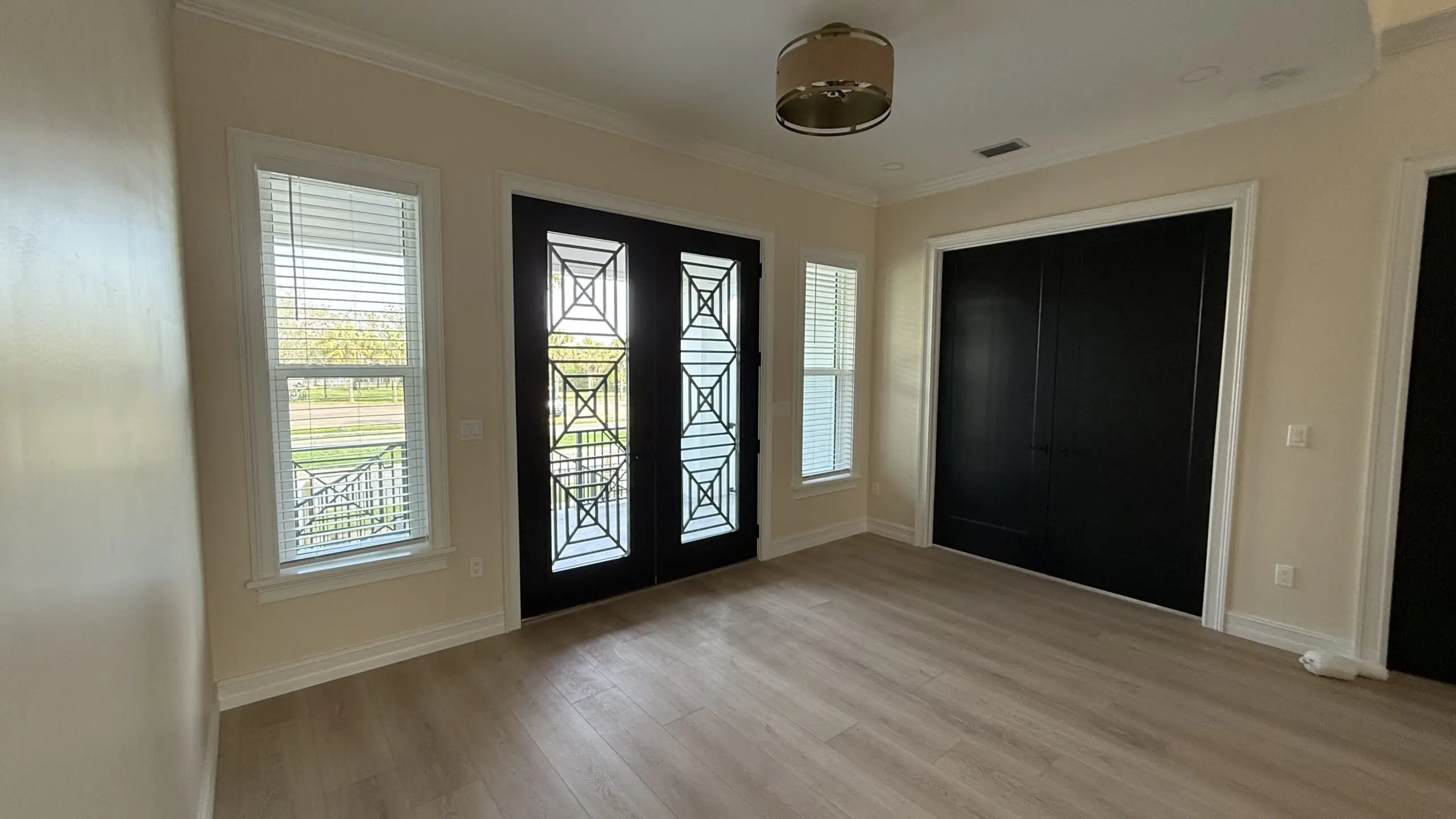 A bright entryway features a double door with decorative metalwork, flanked by two windows with blinds, and a solid black door on the right, all under a ceiling with a pendant light fixture and a vent. A bright entryway features a double door with decorative metalwork, flanked by two windows with blinds, and a solid black door on the right, all under a ceiling with a pendant light fixture and a vent.