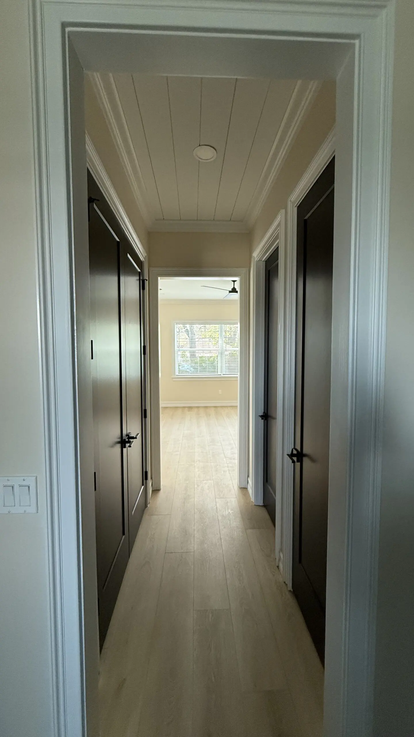 A narrow hallway with light-colored laminate flooring, a white coffered ceiling, and dark-colored doors on either side leads to a brighter room with a window at the far end. A narrow hallway with light-colored laminate flooring, a white coffered ceiling, and dark-colored doors on either side leads to a brighter room with a window at the far end.