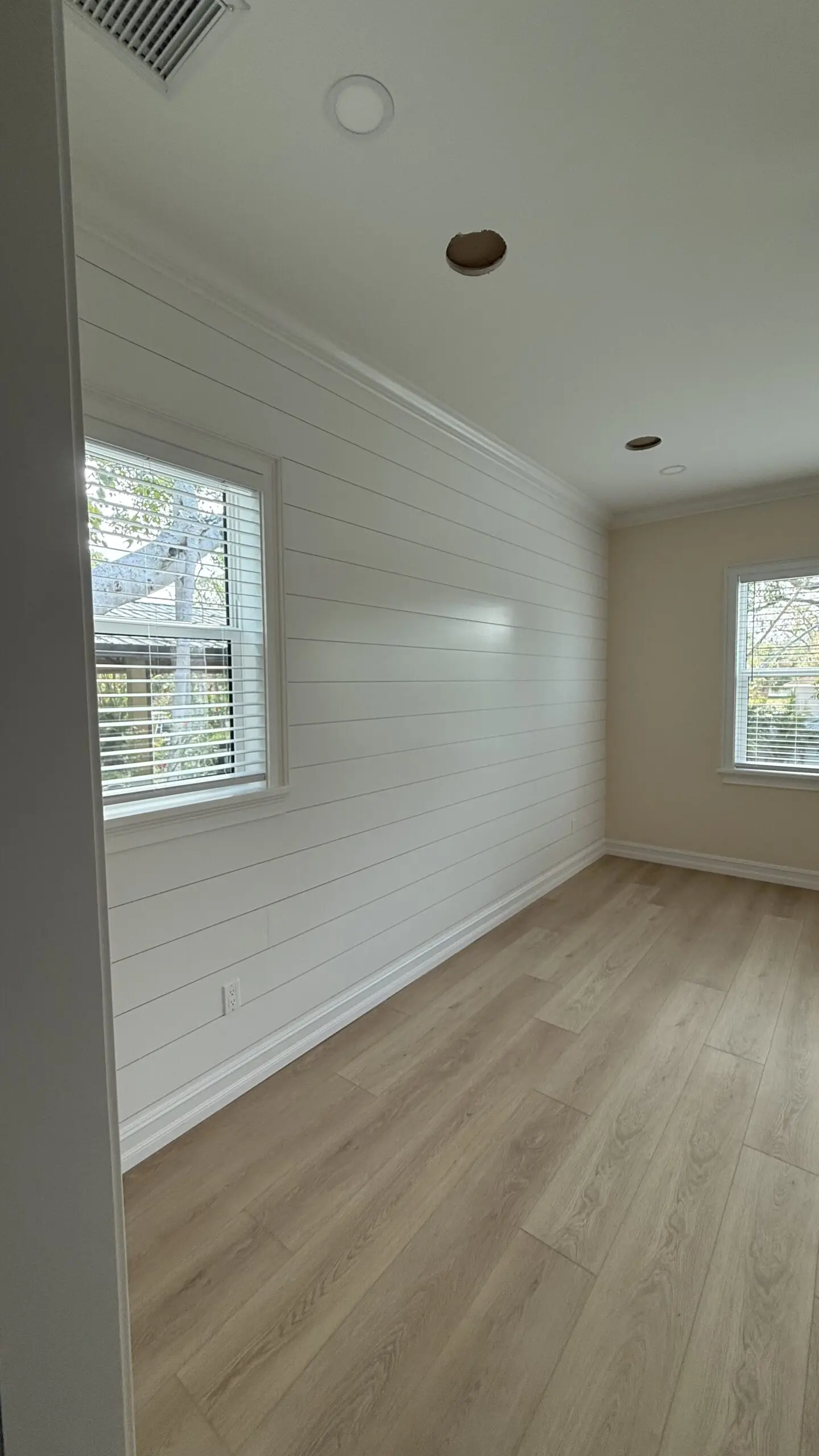 A bright room with white shiplap walls, light wood laminate flooring, two windows with blinds, and recessed lighting in the ceiling, one with an unfinished hole, is shown from a corner angle. A bright room with white shiplap walls, light wood laminate flooring, two windows with blinds, and recessed lighting in the ceiling, one with an unfinished hole, is shown from a corner angle.