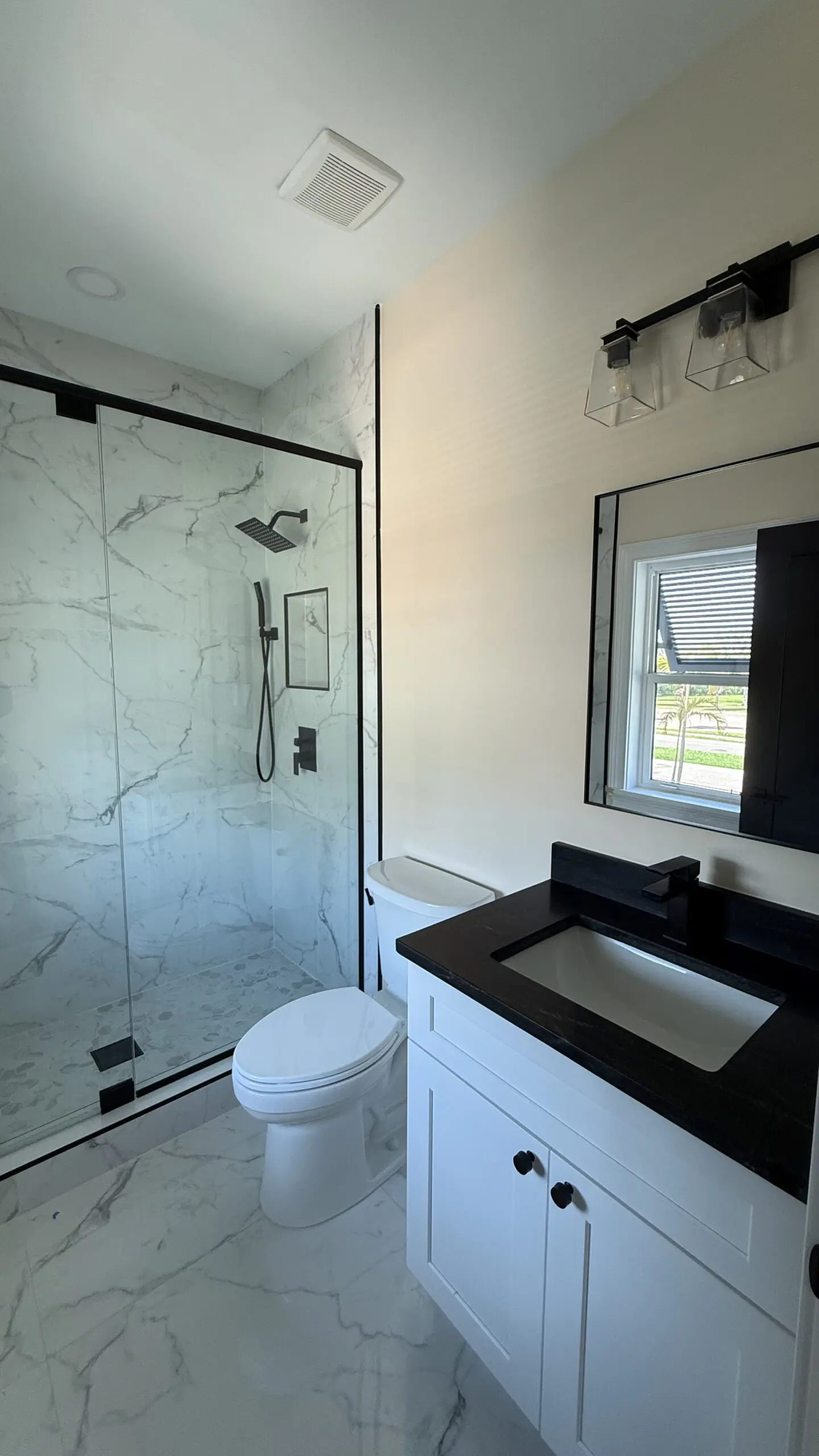 A modern bathroom features a glass-enclosed shower with marble-patterned walls, a white toilet, a white vanity with a black countertop and sink, a framed mirror, and a light fixture with clear glass shades. A modern bathroom features a glass-enclosed shower with marble-patterned walls, a white toilet, a white vanity with a black countertop and sink, a framed mirror, and a light fixture with clear glass shades.