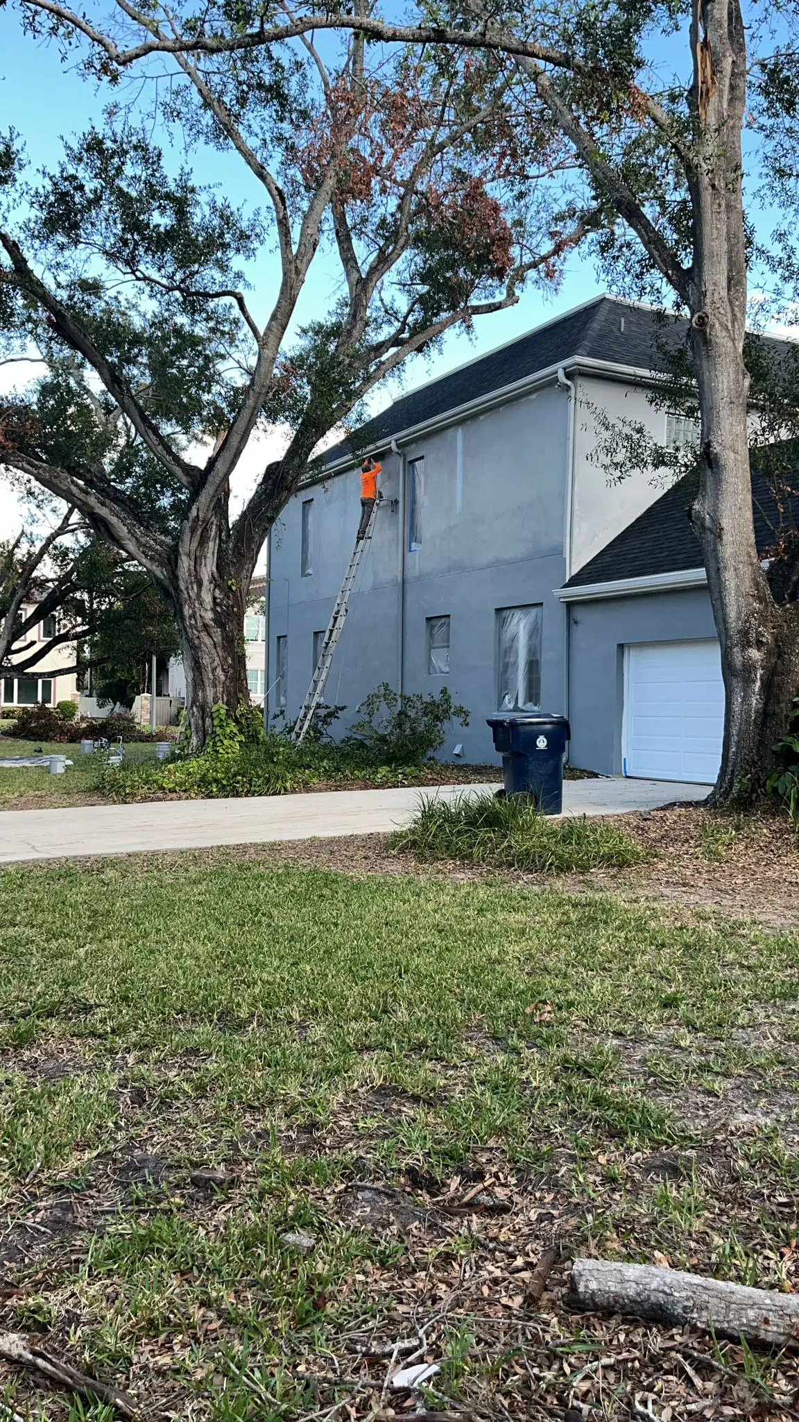 A person wearing an orange vest is on a ladder painting the side of a gray two-story house with a dark gray roof and a white garage door, framed by large trees and a green lawn.