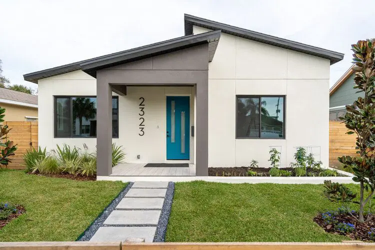 A modern single-story house with a white exterior, gray trim, a teal front door, and a sharply angled roof features the house number "2323" vertically beside the entrance, with a concrete paver pathway leading to the door and landscaping on either side.