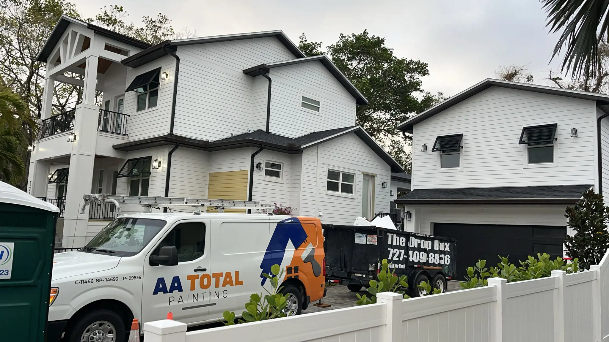 A white van with "AA TOTAL PAINTING" branding is parked in front of two newly painted white houses with black trim and awnings, with a "The Drop Box" trailer and a portable toilet nearby.