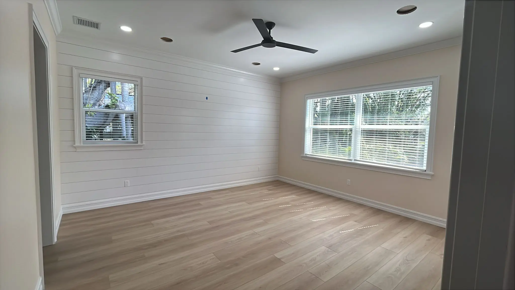A bright room with white shiplap walls, light wood laminate flooring, two windows with blinds, recessed lighting, and a black ceiling fan is viewed from a corner angle. A bright room with white shiplap walls, light wood laminate flooring, two windows with blinds, recessed lighting, and a black ceiling fan is viewed from a corner angle.